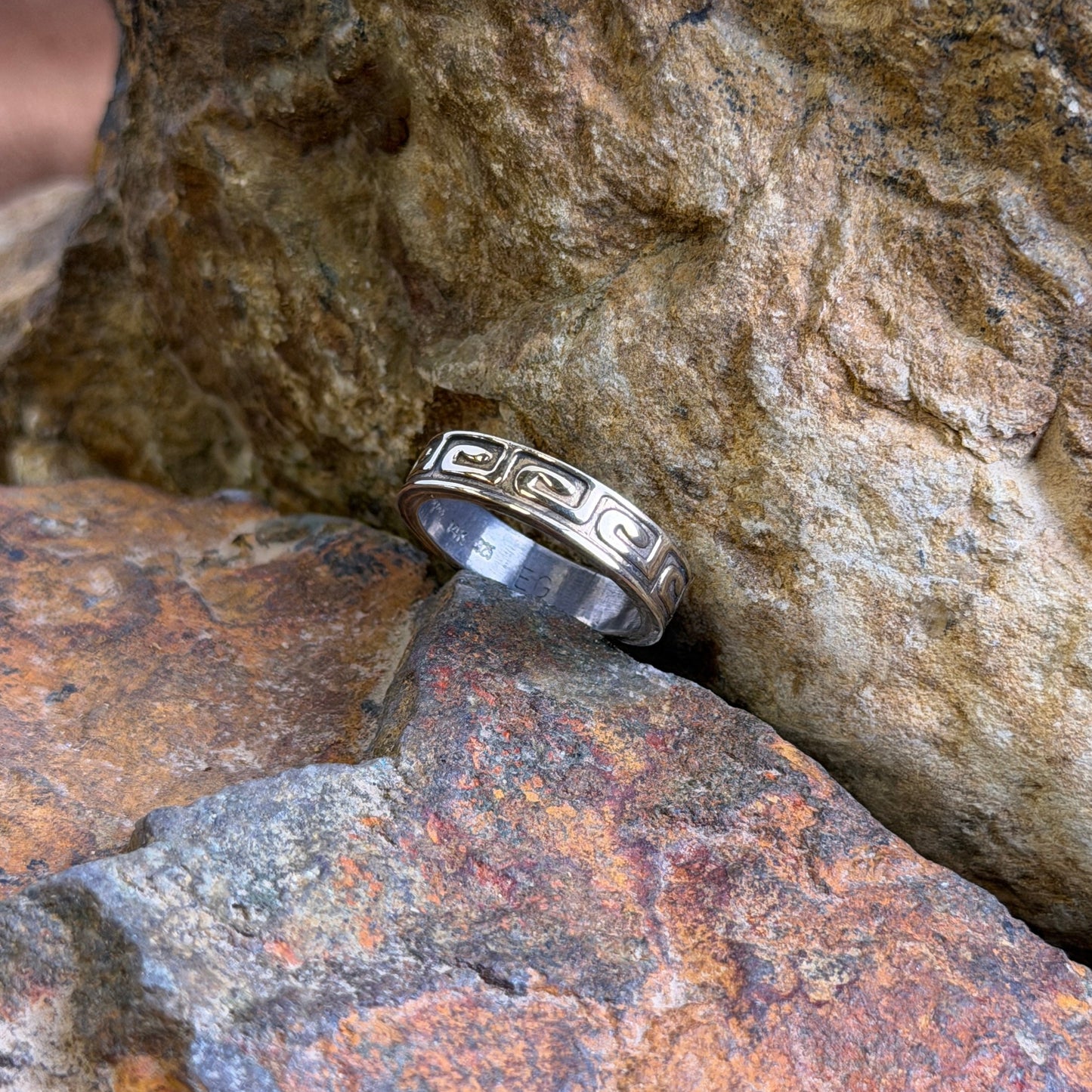 A silver ring with a Greek key pattern, resting on textured orange and brown rocks.