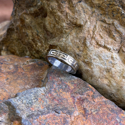 A silver ring with a Greek key pattern, resting on textured orange and brown rocks.