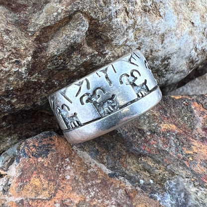 A wide sterling silver band ring with engraved ancient script and animal motifs, resting on textured brown and orange rocks.