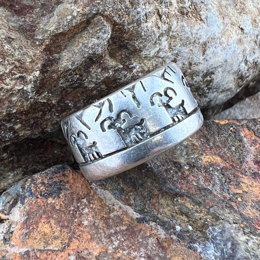 A wide sterling silver band ring with engraved ancient script and animal motifs, resting on textured brown and orange rocks.