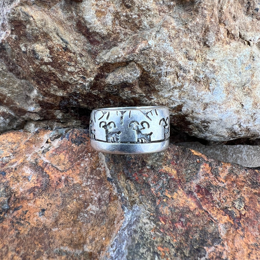 A wide silver band ring with hand-carved animal and geometric engravings, resting on textured orange and brown rocks.