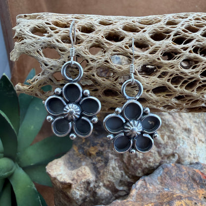 Silver and black floral earrings on a textured stone surface with a plant in the foreground.