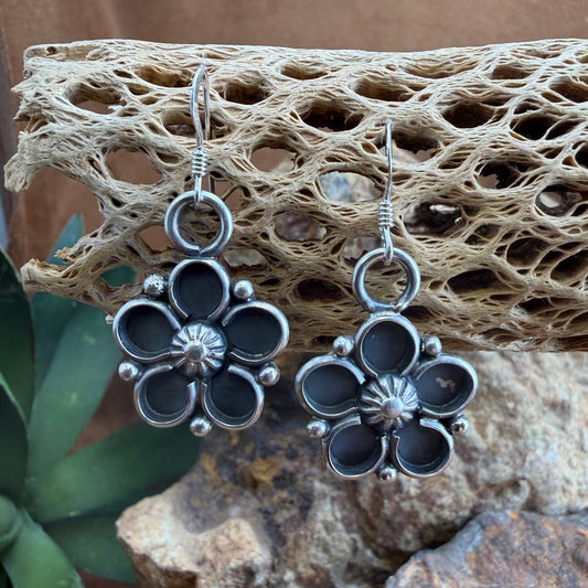 Silver and black floral earrings on a textured stone surface with a plant in the foreground.