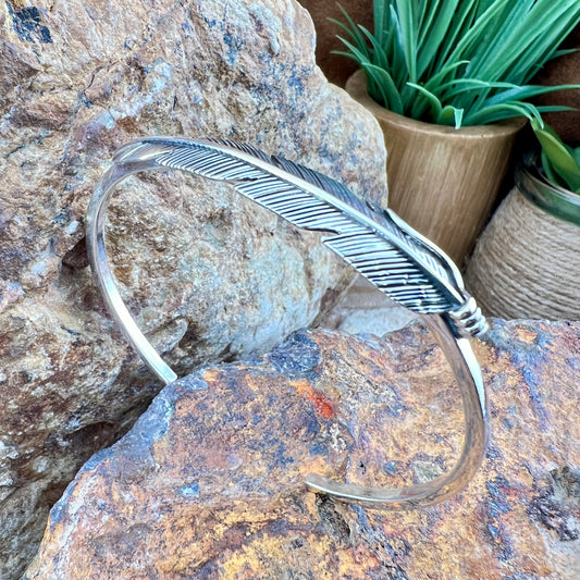 Silver feather-shaped bracelet on a textured stone surface with plants in the background