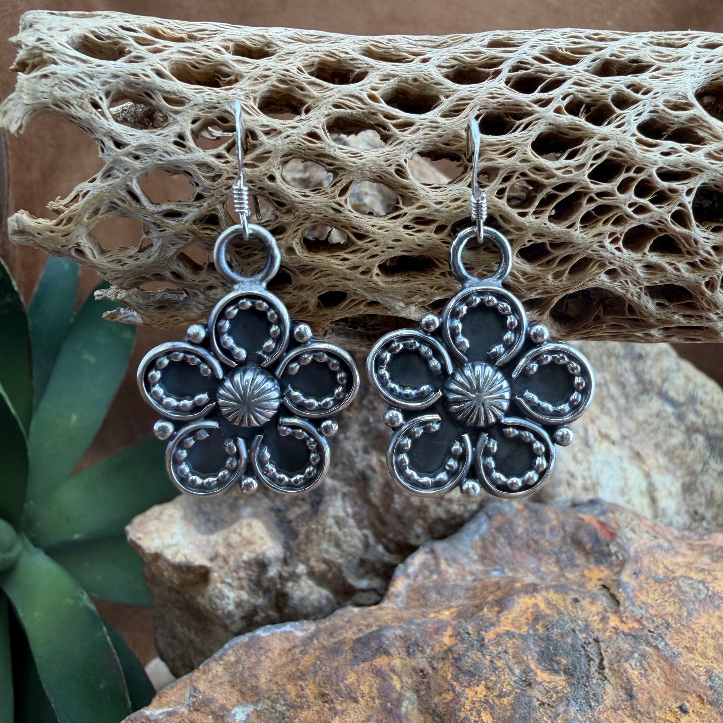 Silver floral earrings on a textured stone surface with a plant in the background