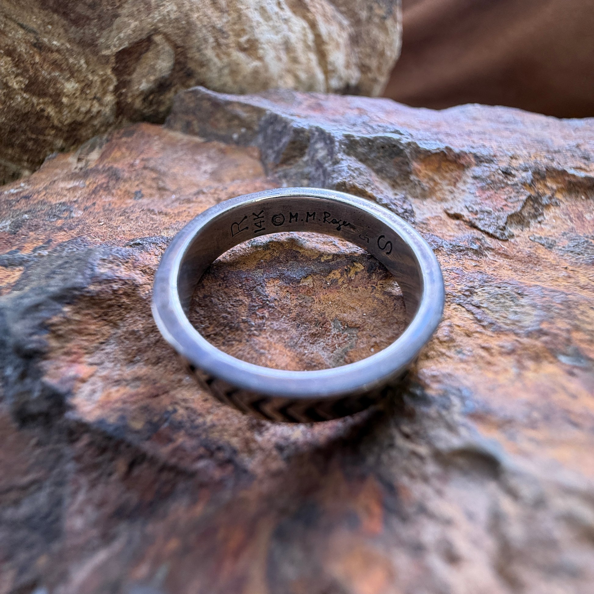 A sterling silver band ring with a textured lower half and engraved initials inside, resting on a rustic, textured brown rock.