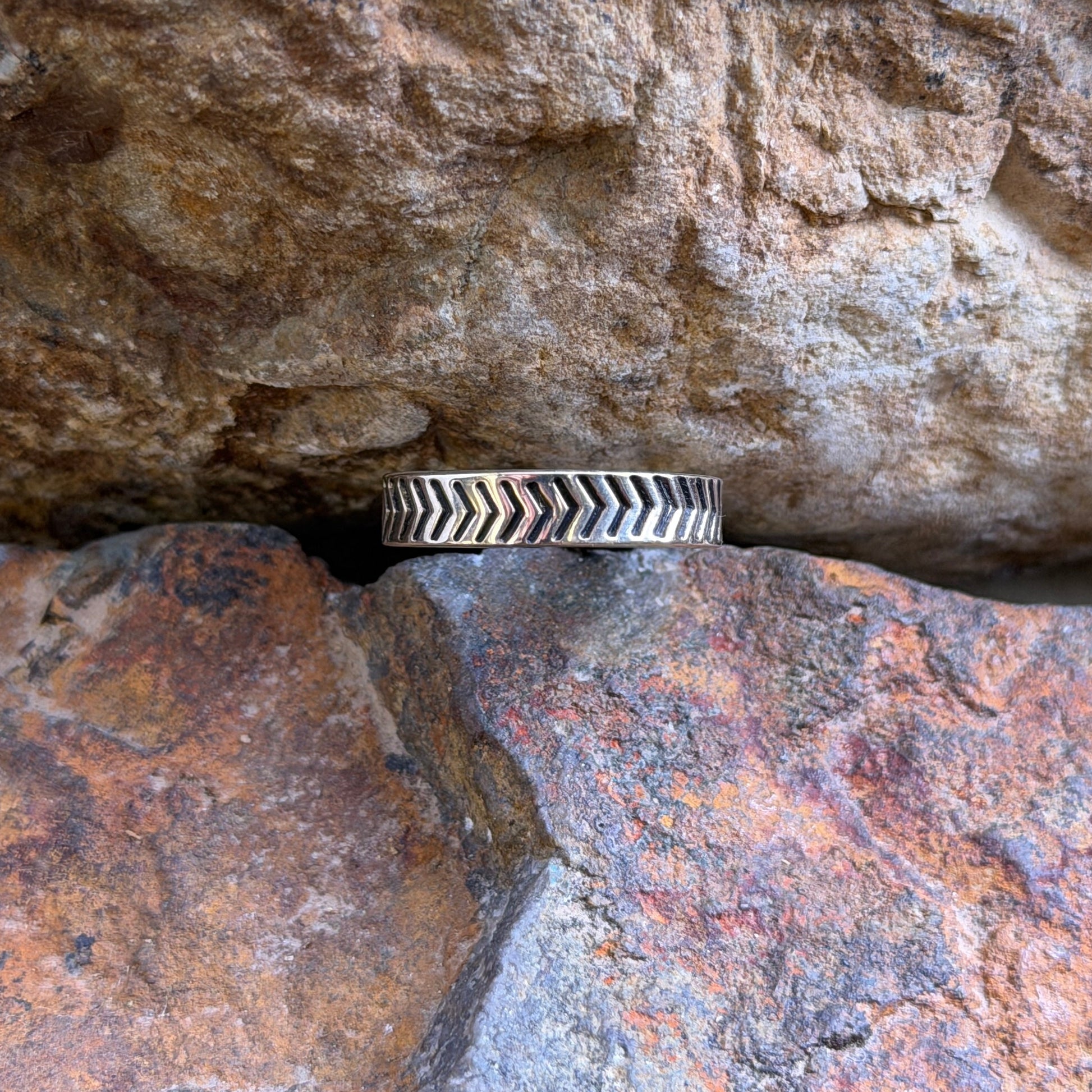 A silver ring with a chevron or zigzag pattern, resting on textured orange and brown rocks.