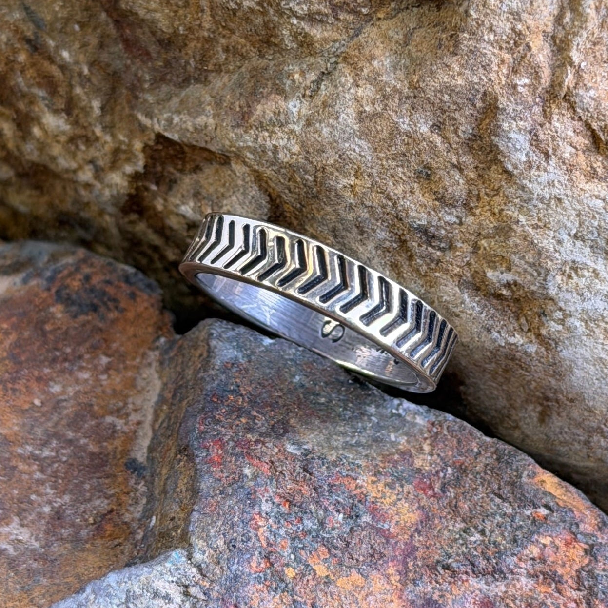 A silver ring with a chevron or zigzag patterned band, resting on textured orange and brown rocks.