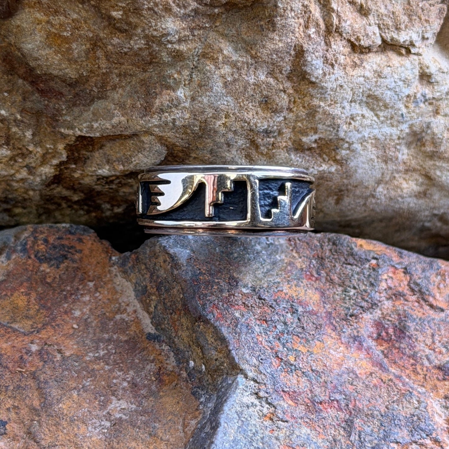A silver ring with black inlay and geometric cutout details, resting on a textured orange-brown rock surface.