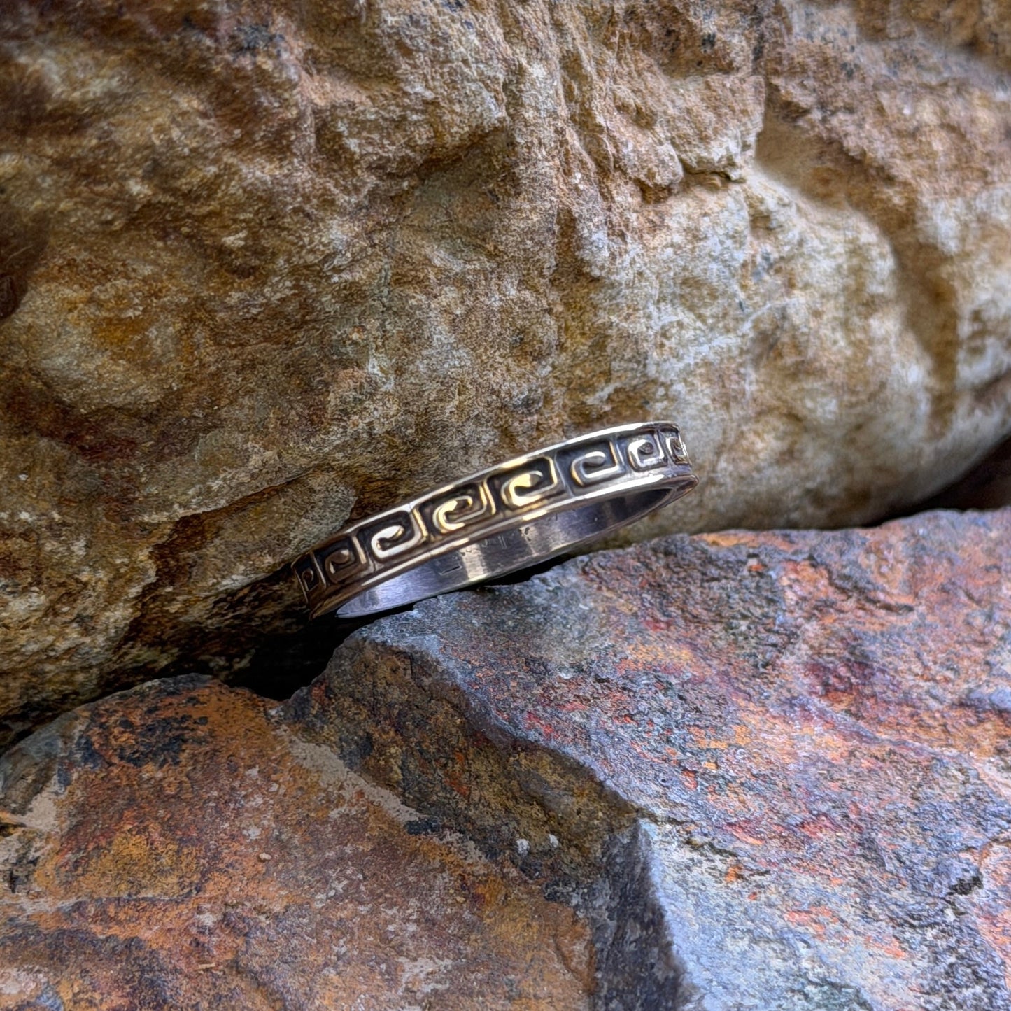 A silver band ring with a Greek key pattern engraved on the side, resting between two textured natural rocks.