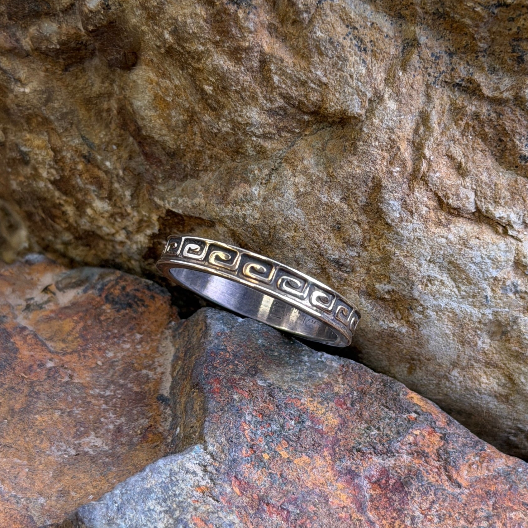 A silver band ring with a repeating geometric key motif, resting on textured brown and orange rocks.