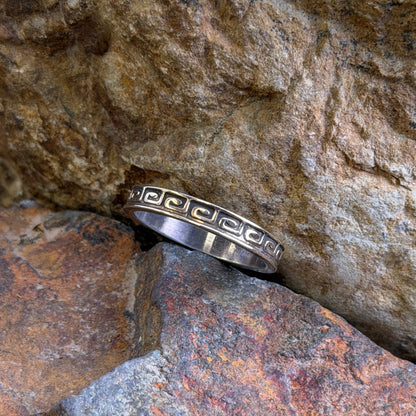 A silver band ring with a repeating geometric key motif, resting on textured brown and orange rocks.