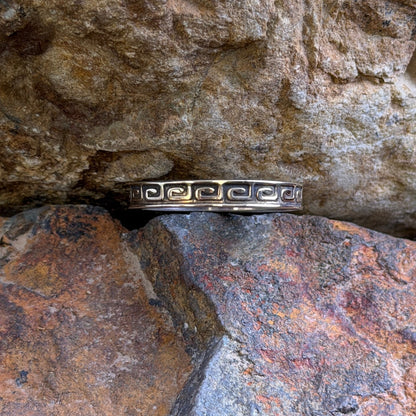 A silver ring with a repeating geometric pattern, resting on a textured brown and orange rock surface.