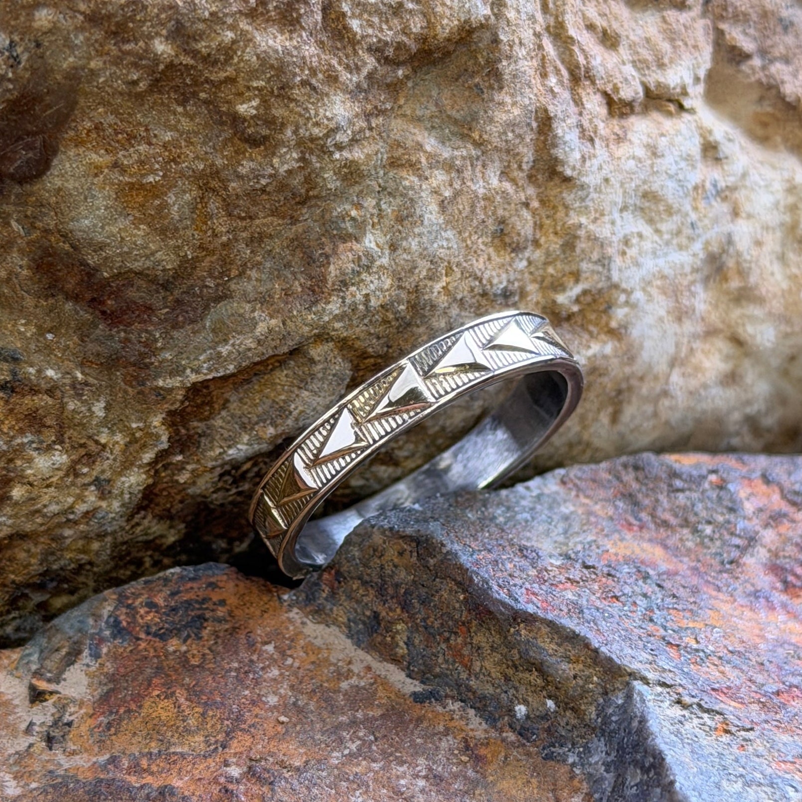 A silver-toned band ring with engraved geometric chevron patterns, resting on textured brown and orange rocks.