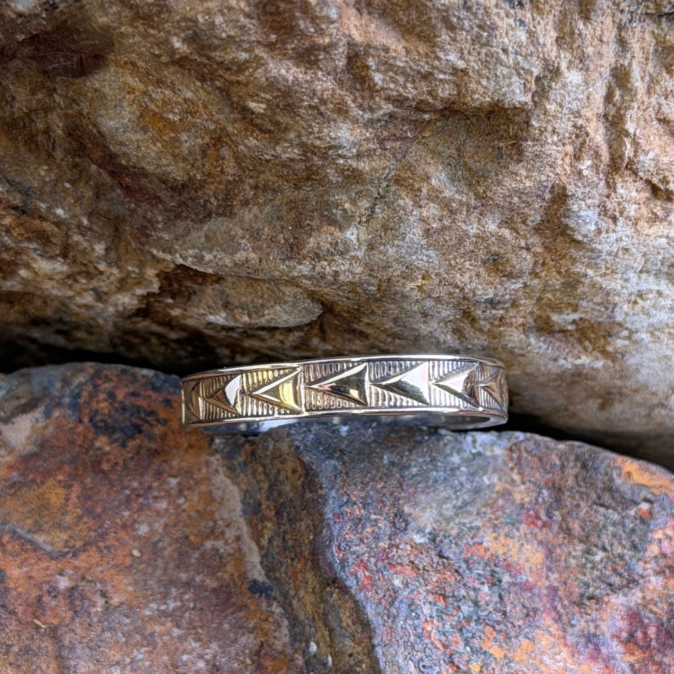 A gold band ring with engraved geometric chevron patterns, resting on a textured orange-brown rock against a larger brown rock background.