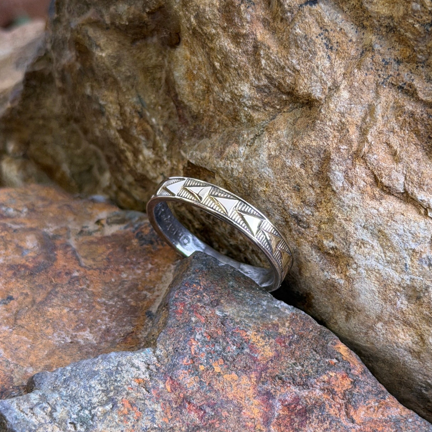 A silver band ring with a geometric chevron pattern, resting on textured orange and brown rocks.