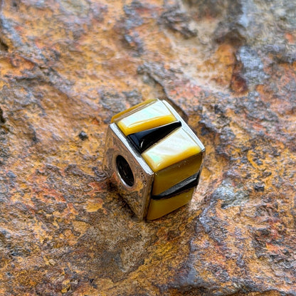 A small cube-shaped pendant charm with alternating yellow and black striped segments, resting on a textured, rusted brown rock surface.