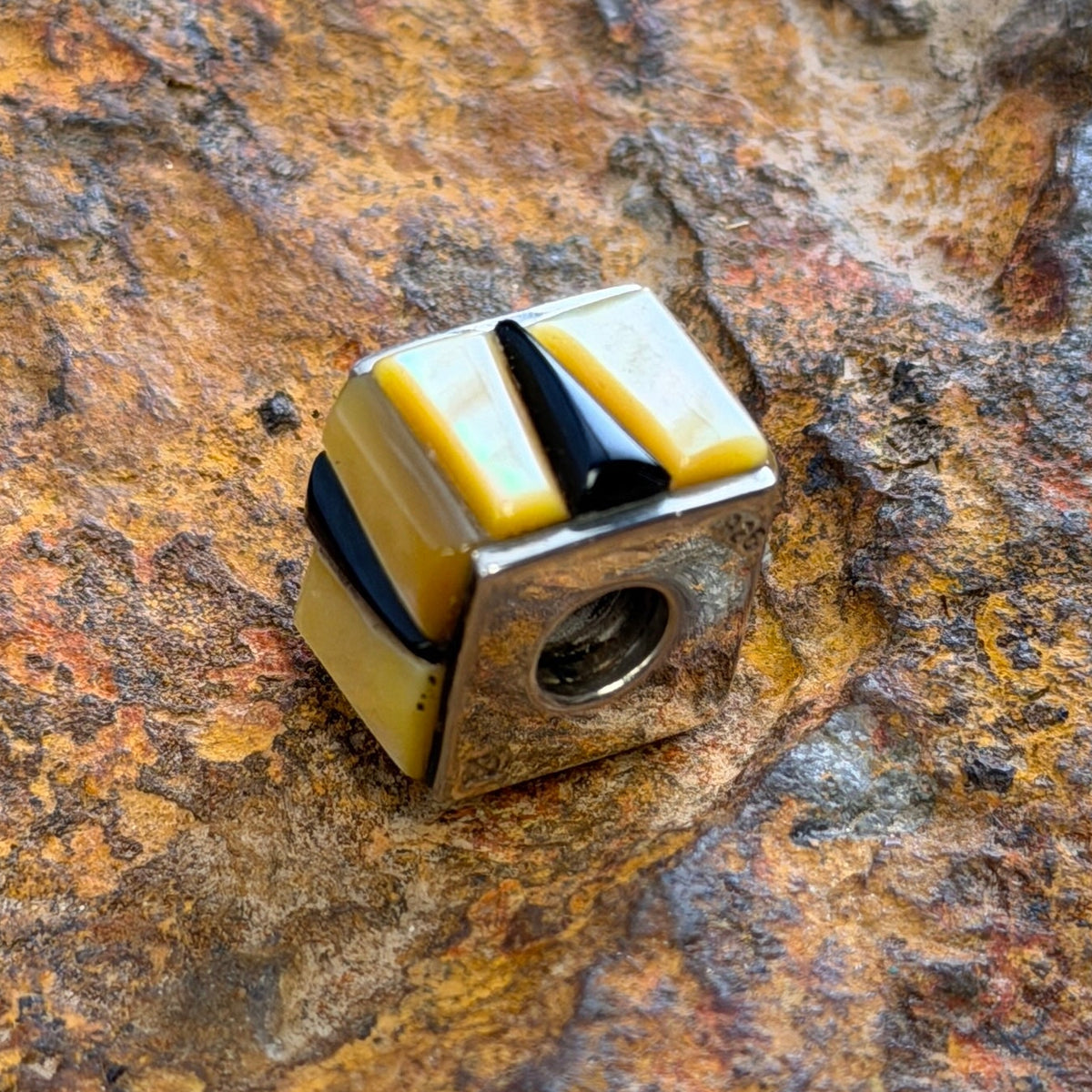 A small cube-shaped pendant charm with alternating yellow and black striped segments, resting on a textured, rusted brown rock surface.