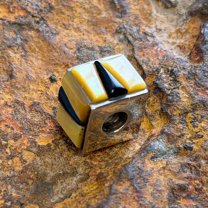 A small cube-shaped pendant charm with alternating yellow and black striped segments, resting on a textured, rusted brown rock surface.