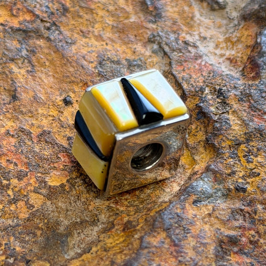A small cube-shaped pendant charm with alternating yellow and black striped segments, resting on a textured, rusted brown rock surface.