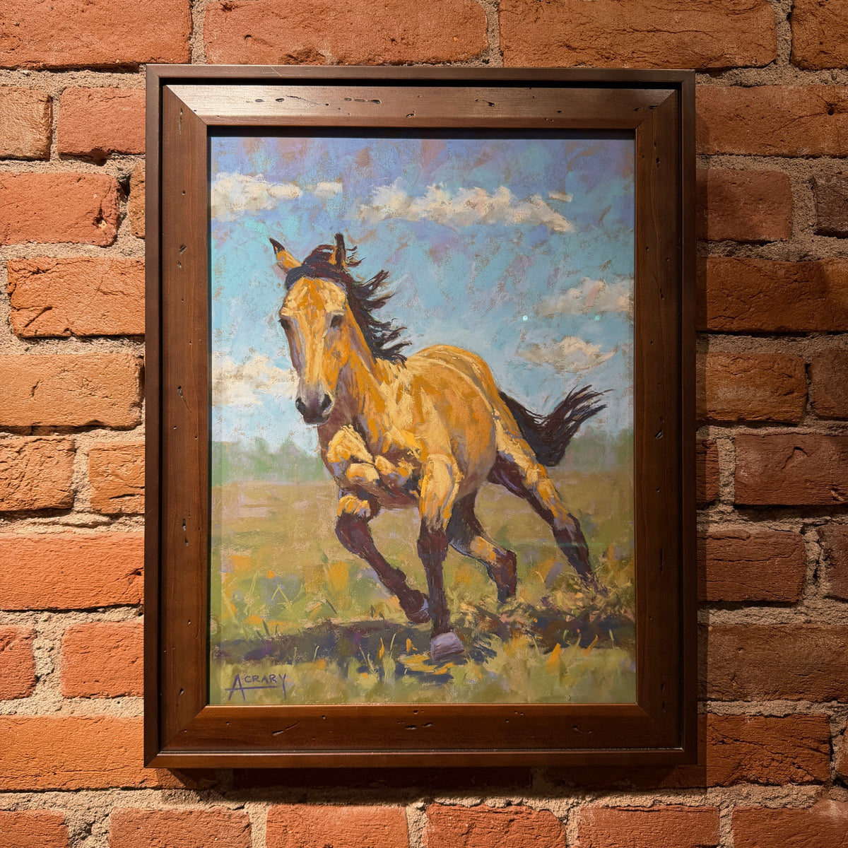 A framed painting of a galloping horse in a grassy field under a blue sky, mounted on a red brick wall.