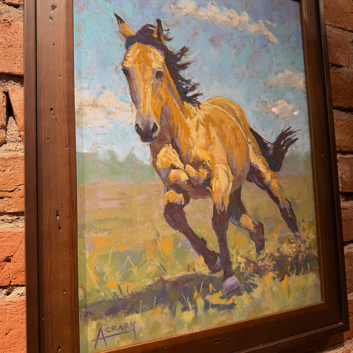A framed painting of a galloping horse in a grassy field under a blue sky, mounted on a brick wall. The painting features expressive brushstrokes in tan, brown, black, green, and blue, and is housed in a dark wood frame with a visible artist signature at the bottom.