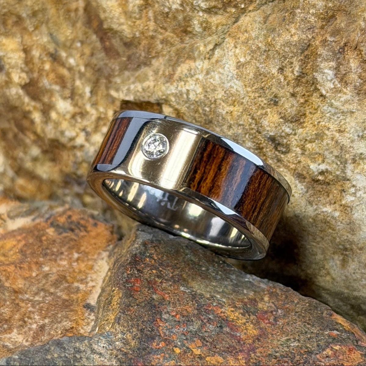 A ring with a dark brown wood inlay and a small clear diamond, resting on textured orange-brown rocks outdoors
