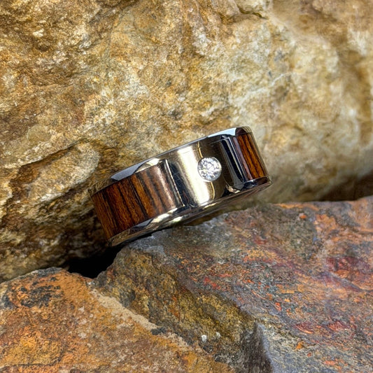 A ring with a dark brown wood inlay and a small clear diamond, resting on textured orange-brown rocks outdoors