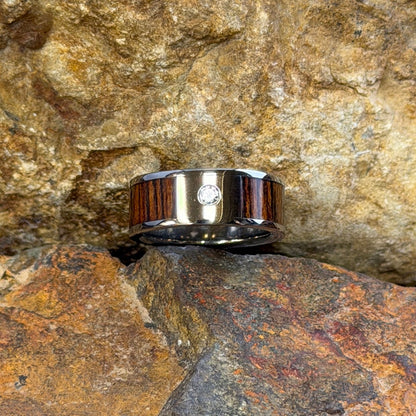 A ring with a dark brown wood inlay and a small clear diamond, resting on textured orange-brown rocks outdoors