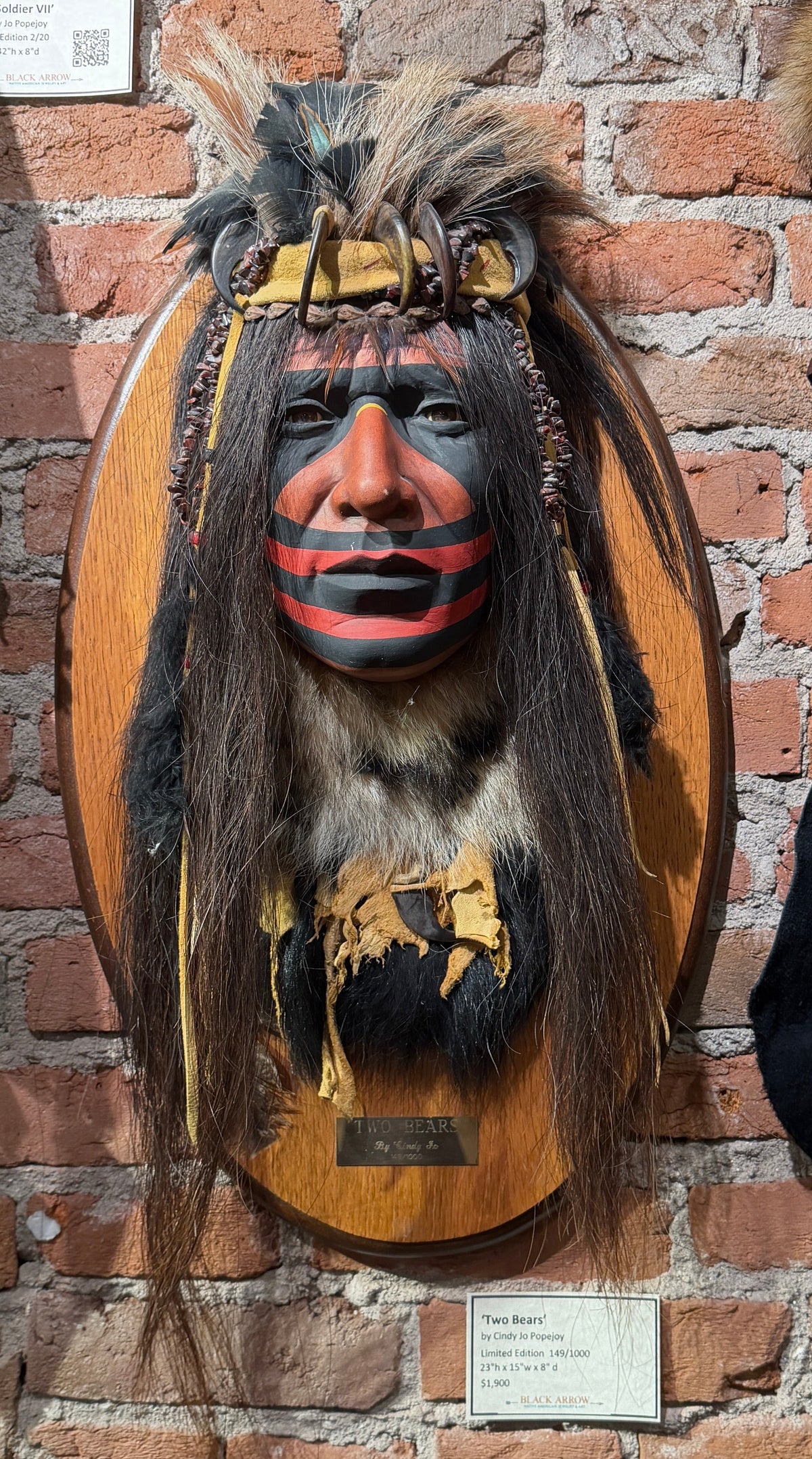 A mounted Native American-style headpiece with a painted face, long dark hair, and feather details attached to an oval wooden plaque, displayed against a red brick wall with other exhibits partially visible at the sides.