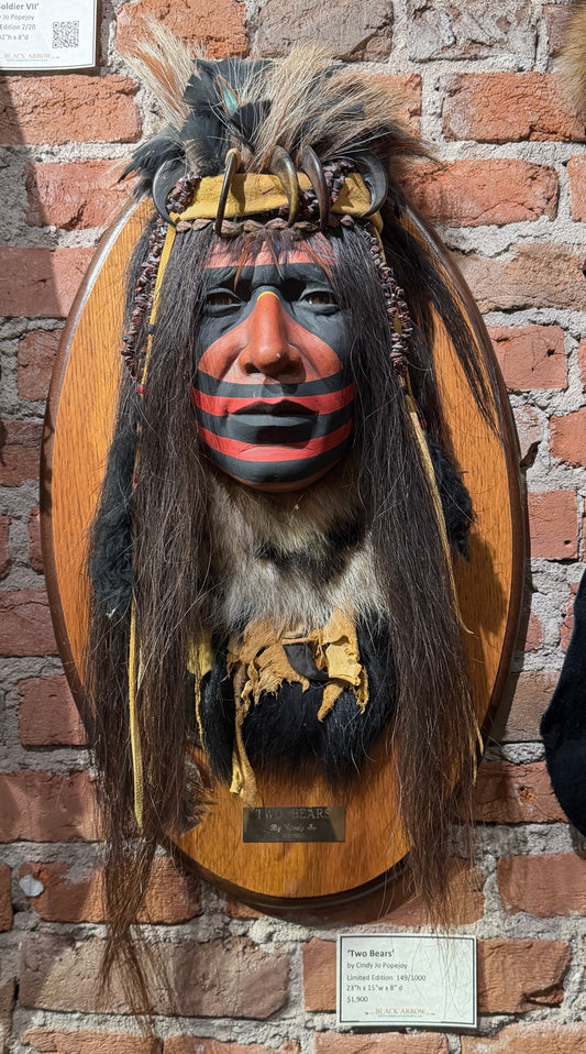 A mounted Native American-style headpiece with a painted face, long dark hair, and feather details attached to an oval wooden plaque, displayed against a red brick wall with other exhibits partially visible at the sides.