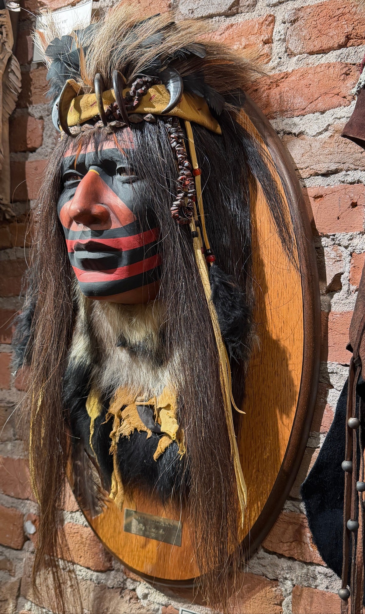 A carved Native American-style face mask with long dark hair, a tan headband, and red and black facial paint, mounted on a round wooden plaque against a brick wall. Traditional garments hang nearby on the left and right.