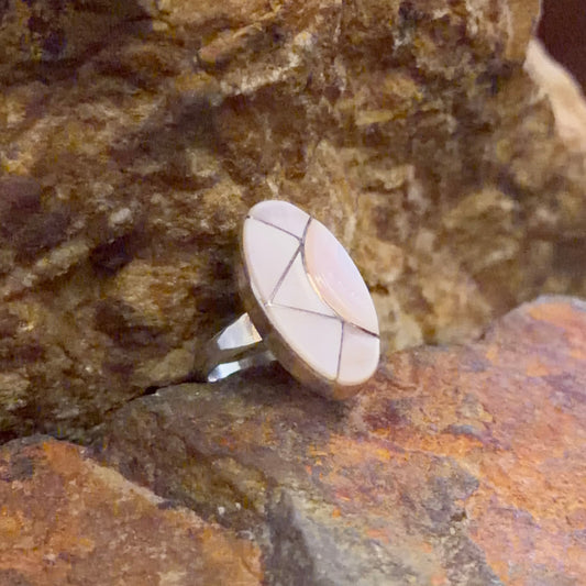 A silver ring with a circular inlay of light pinkish-white inlay work. nestled between textured brown and orange rocks.