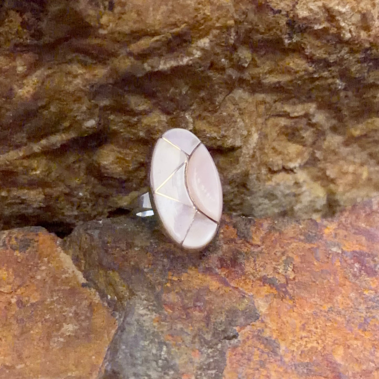 A close-up of a ring with a round, light pink stone set in a silver-toned band, nestled between textured brown and orange rocks.