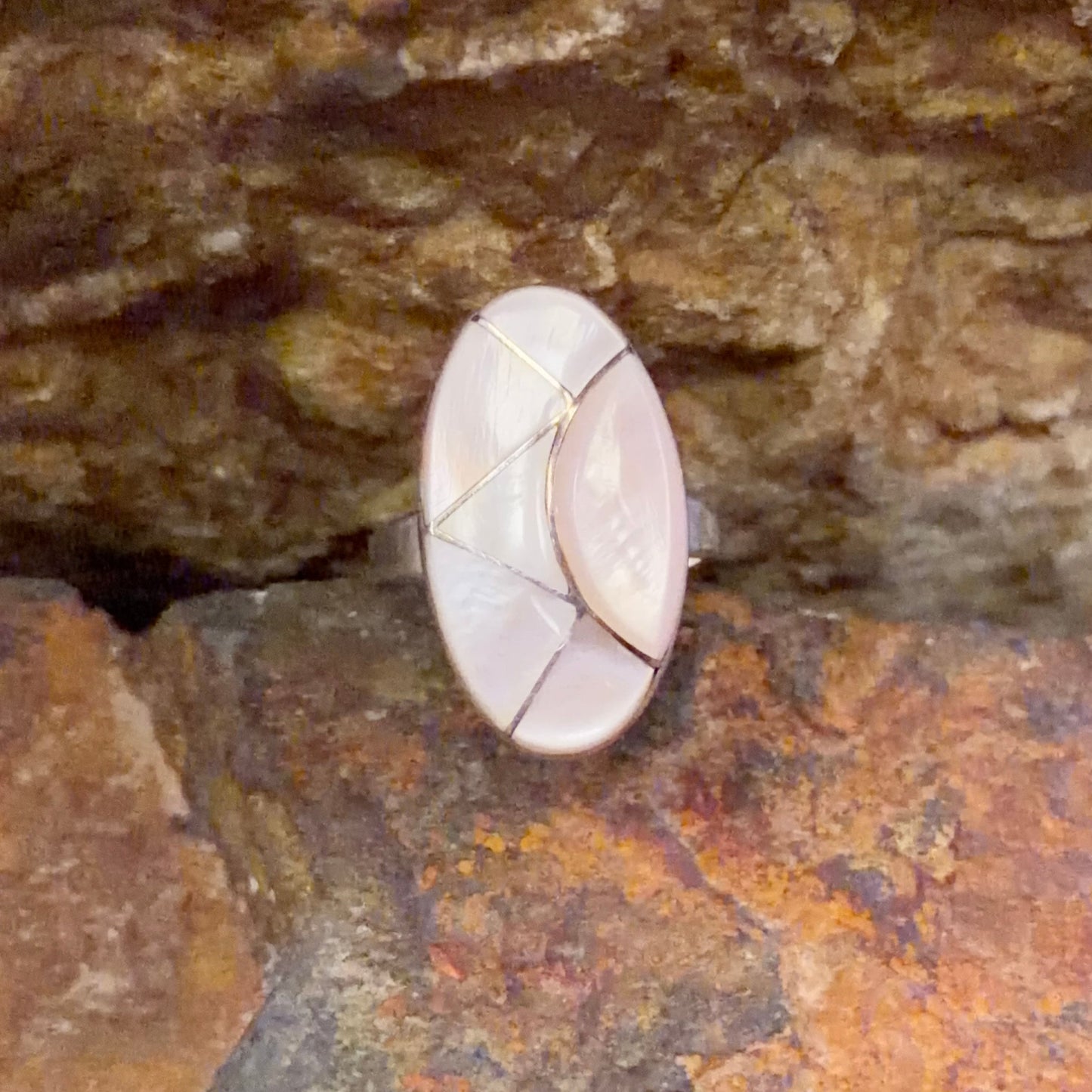 Oval ring face inlaid with light pink segments. nestled in-between a rocky surface