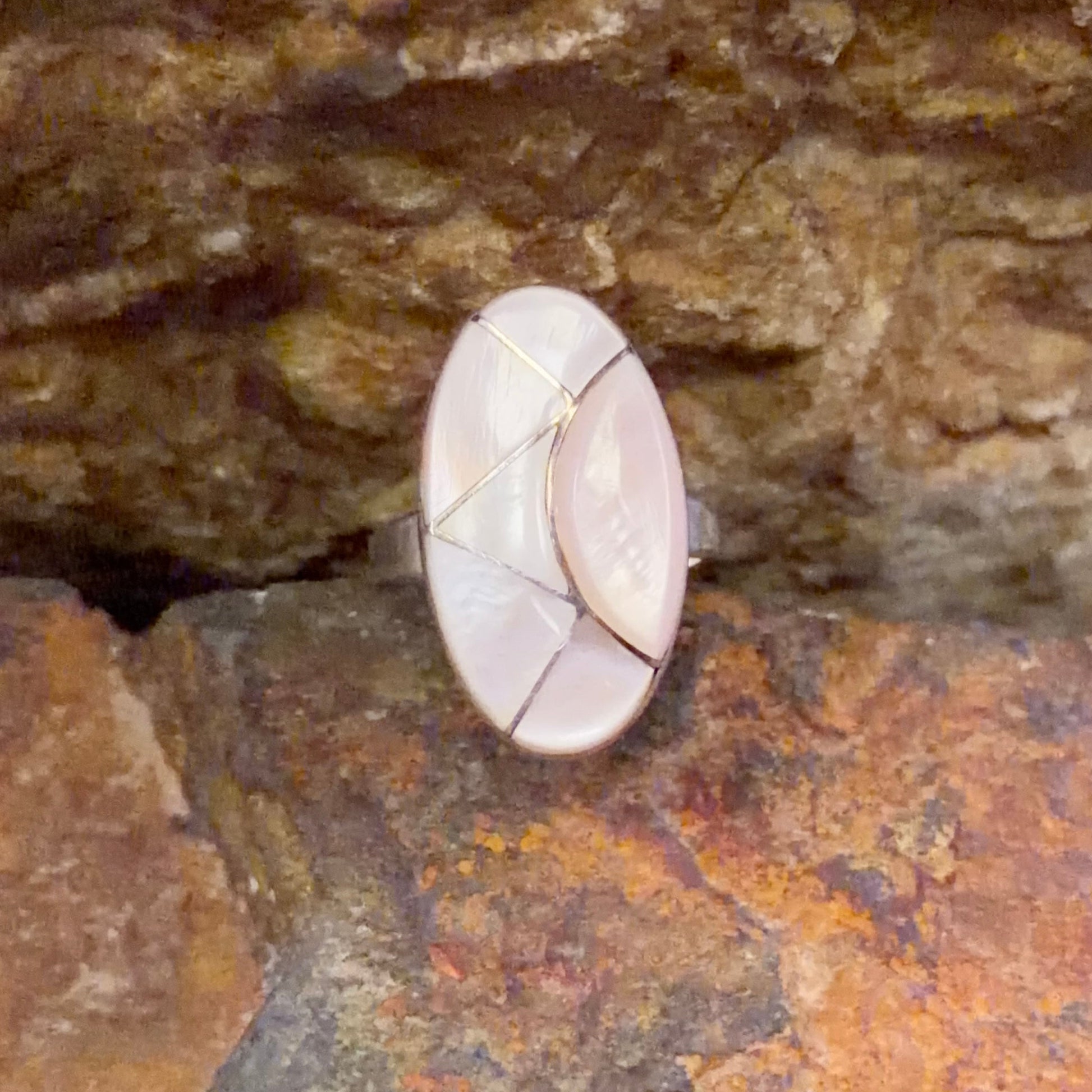 Oval ring face inlaid with light pink segments. nestled in-between a rocky surface