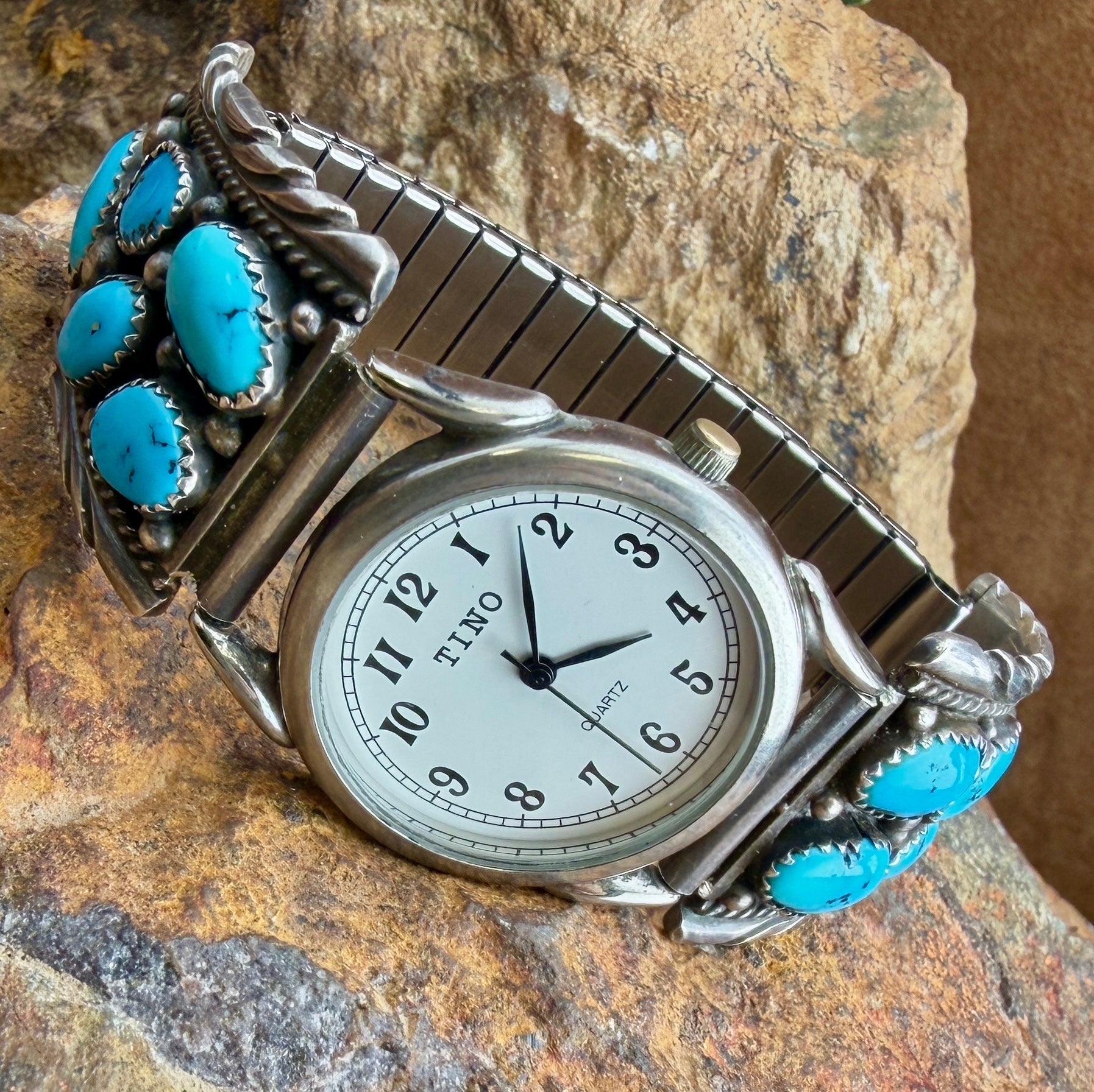 A silver-toned metal watch with a curved bracelet featuring multiple round turquoise stones, resting on a textured rock surface. The watch face is white with black Arabic numerals and hands.