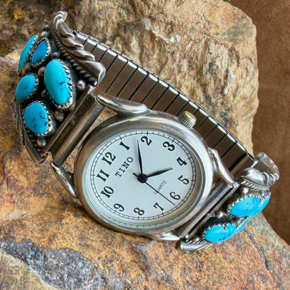 A silver-toned metal watch with a curved bracelet featuring multiple round turquoise stones, resting on a textured rock surface. The watch face is white with black Arabic numerals and hands.