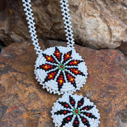 A close-up of a beaded necklace laid on rough brown rocks. The necklace has a white beaded chain and two round white bead pendants with a geometric floral/starburst pattern in black, red, yellow, and green.