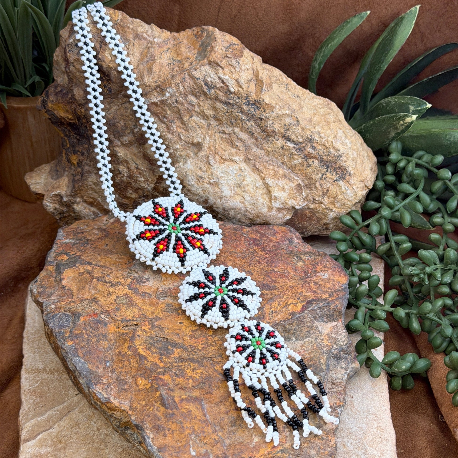 A beaded necklace displayed on a textured rock, featuring a white seed bead chain, three circular starburst bead motifs in red, green, and black, and a dangling tassel section with black and white bead fringe.