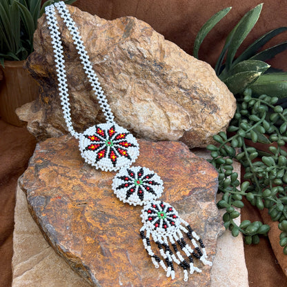 A beaded necklace displayed on a textured rock, featuring a white seed bead chain, three circular starburst bead motifs in red, green, and black, and a dangling tassel section with black and white bead fringe.