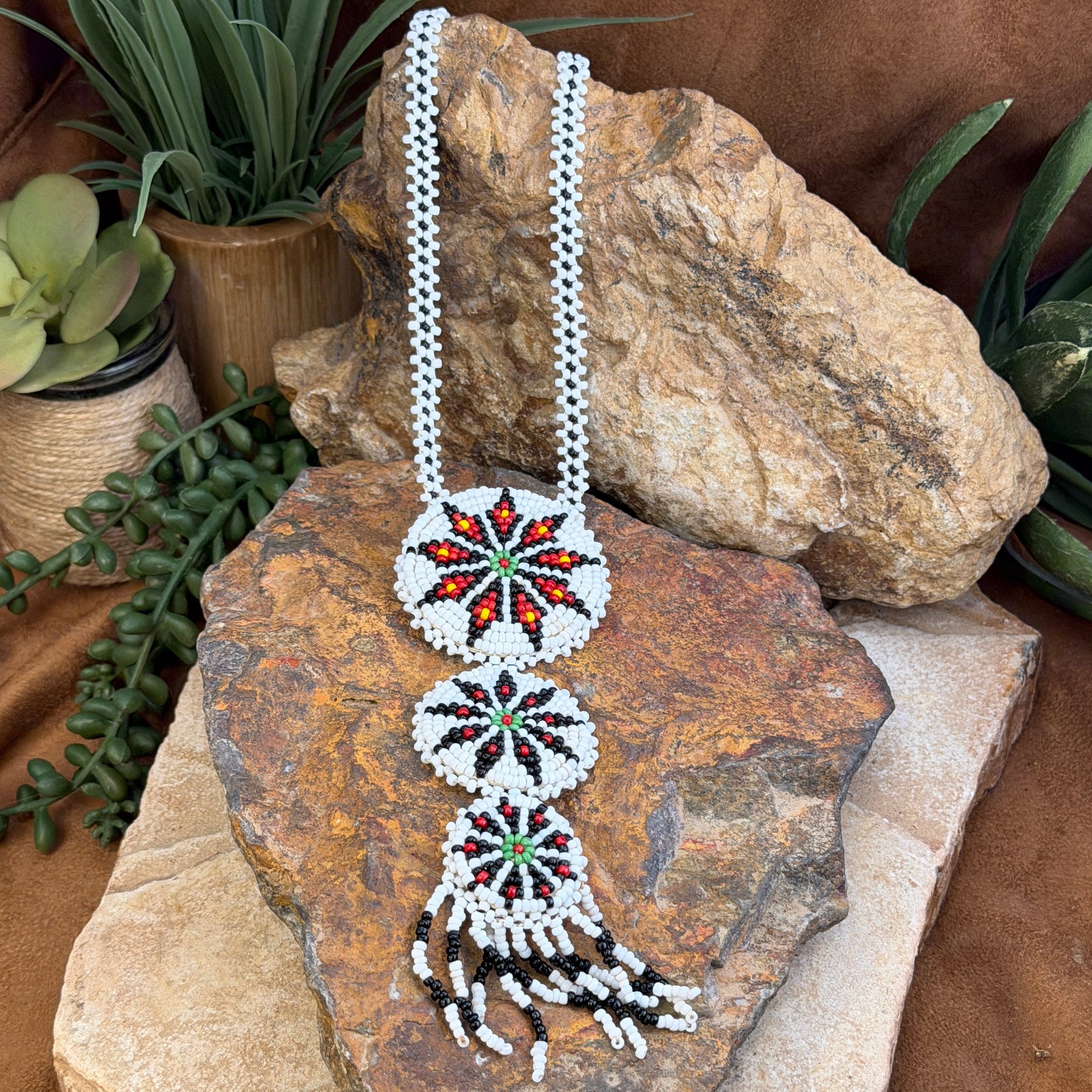 A beaded headband with three circular starburst motifs and a feather-like end detail, displayed on rustic rocks with plants and twine as styled props.