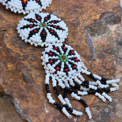 Close-up of a beaded jewelry piece laid on a textured brown rock surface. The visible portion shows circular white, black, and red bead motifs with long dangling fringe in black and white beads.