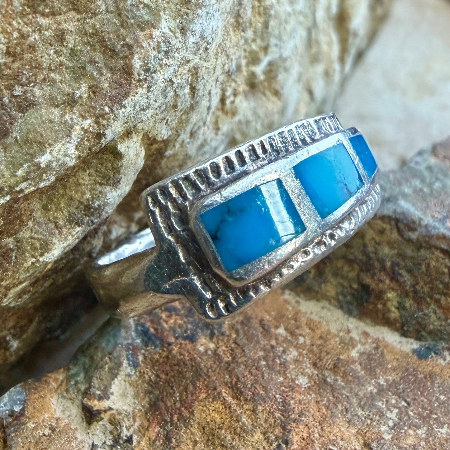 A silver ring with  rectangular blue turquoise inlays, resting on a textured brown and gray rock surface.