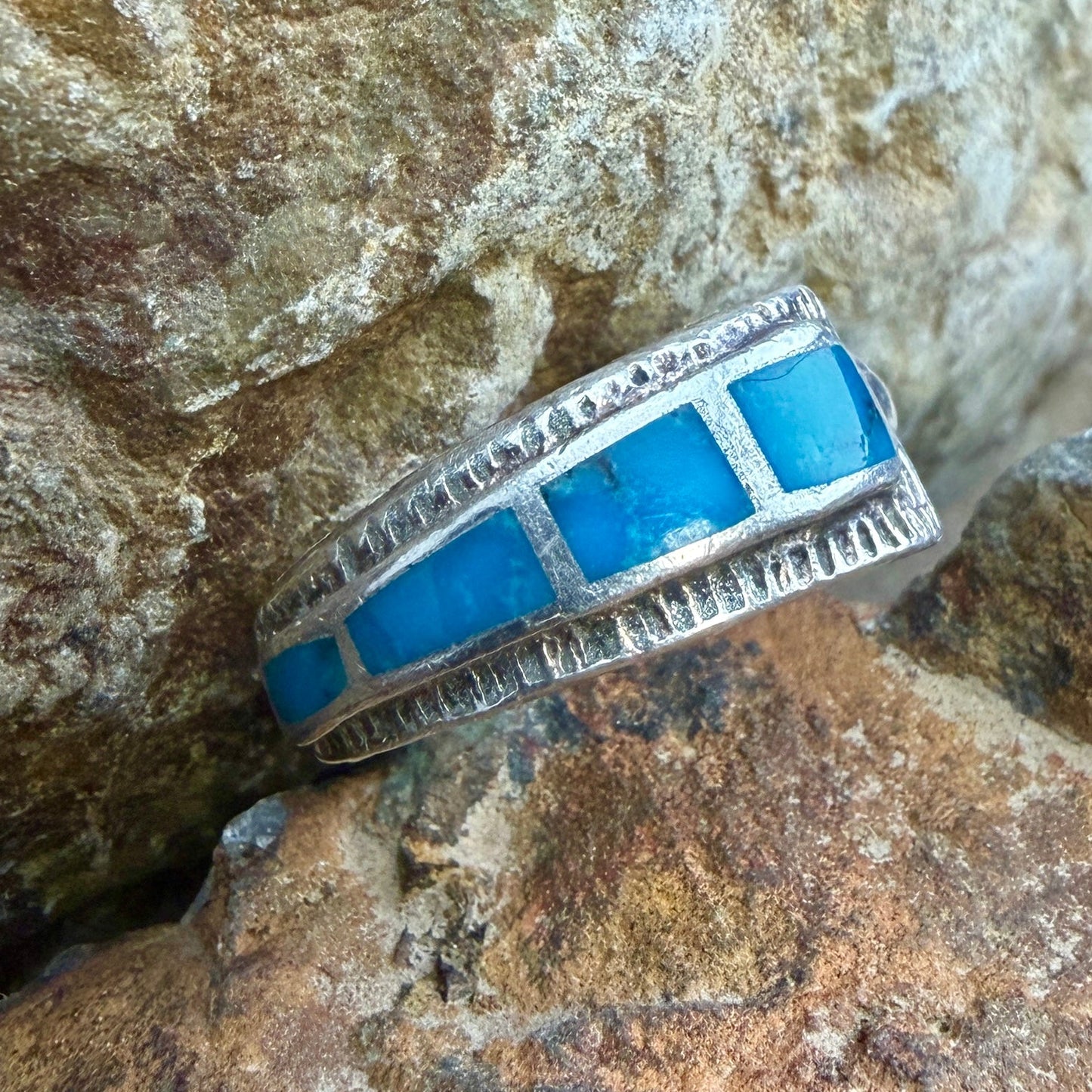 A silver ring with rectangular blue inlay stones, photographed resting on rough natural rocks.