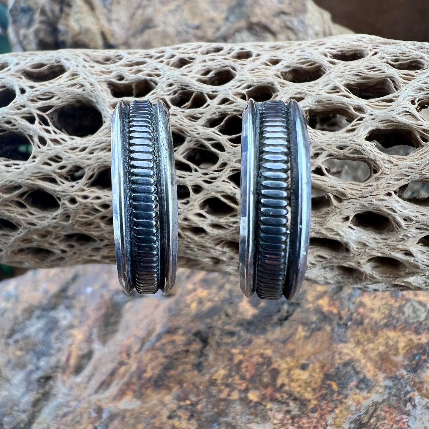 A pair of silver-toned bangles with dark, ribbed inlay bands, displayed on a textured, porous beige stone prop over a natural rock background.