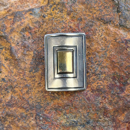 A small rectangular metal jewelry piece with layered silver-toned and gold-toned plates, displayed on a textured orange-brown rock surface.