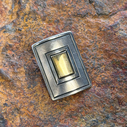 A rectangular silver-toned ring with a gold-toned center, displayed on a textured brown and orange rock surface.