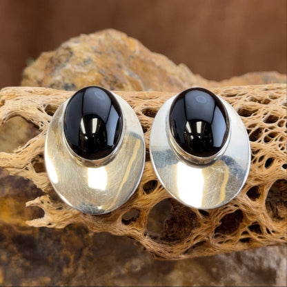 A pair of silver-toned stud earrings with oval black cabochon stones, displayed on a textured natural wood branch against a warm brown background.