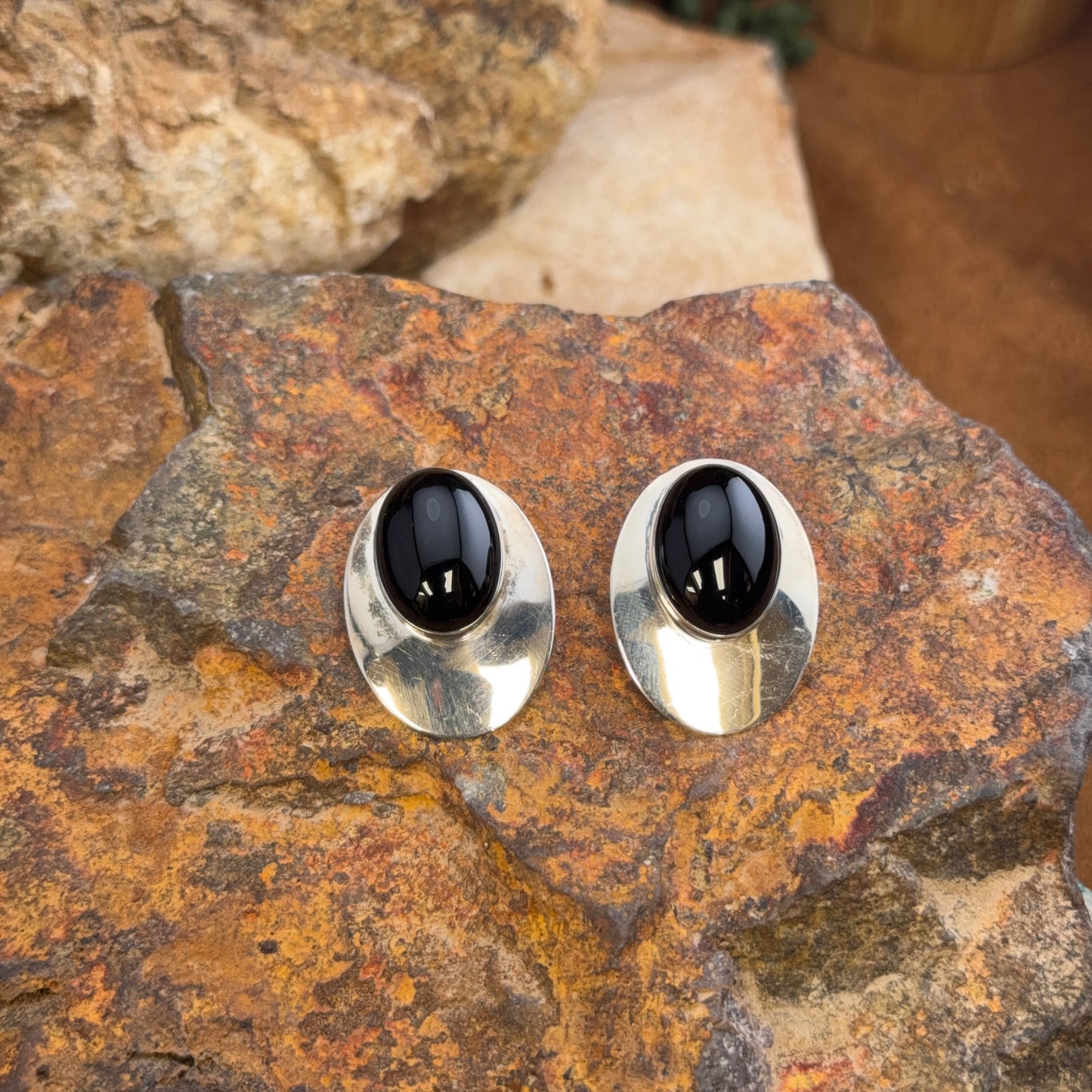 A pair of oval stud earrings with polished silver-toned metal settings and smooth black cabochon centers, displayed on a textured brown and orange rock surface.